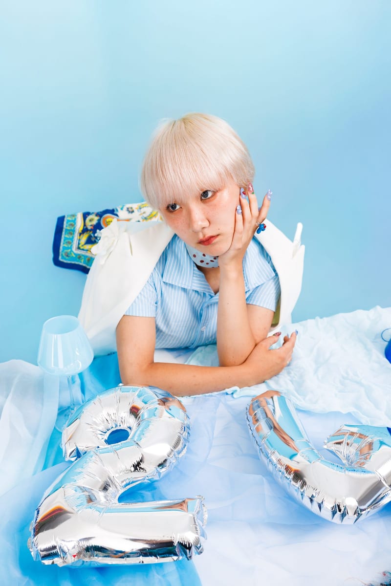 Leaning on a table among silver number balloons, a person rests their chin on their hand