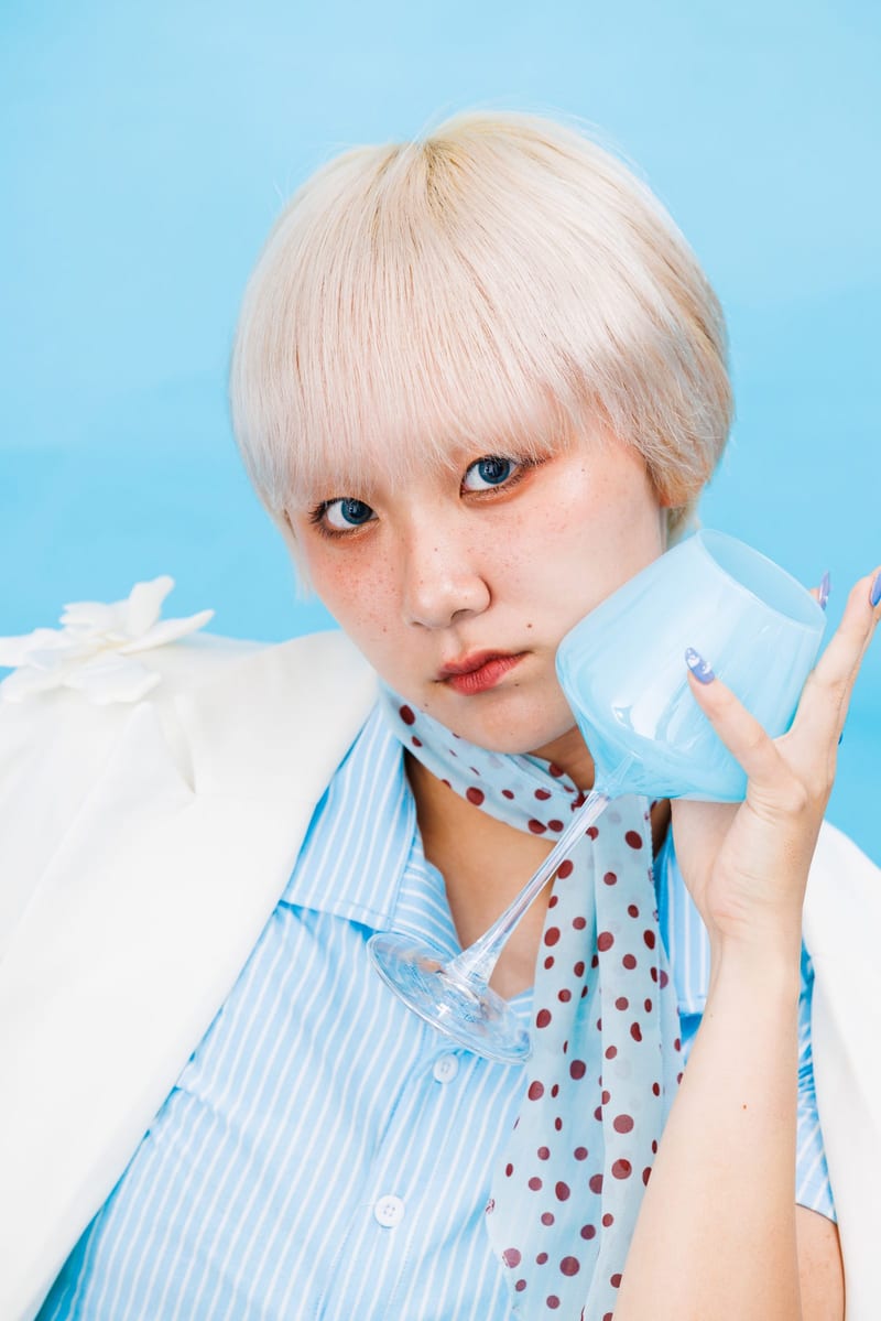 Close up, a person in a polka dot scarf presses a pale blue cup to their cheek