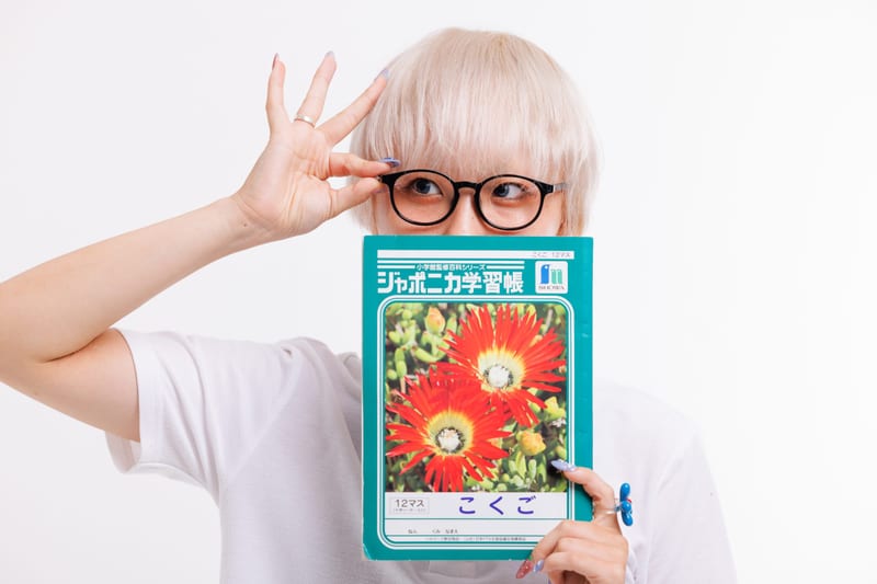 On a white background, a person in glasses holds up a school notebook with a red flower on its cover